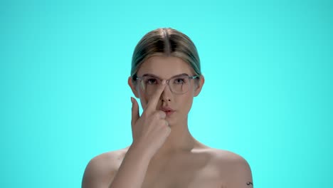 Medium-static-shot-of-a-pretty-glasses-wearing-woman-looking-expectantly-into-the-camera-while-adjusting-her-glasses-in-her-pretty-face-against-turquoise-background-in-slow-motion