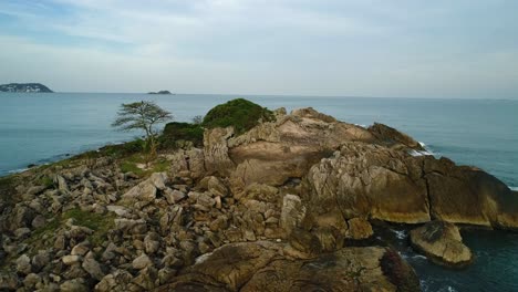 Plane-with-drone-over-a-small-stone-island-in-calm-and-peaceful-sea,-with-waves-crashing-on-the-rocks,-guarujá,-Brazil-beach