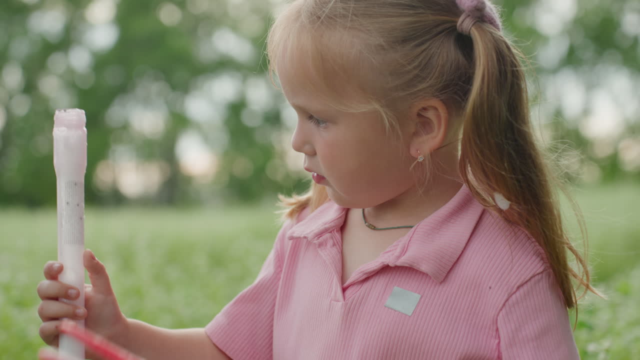 Niña blanca al aire libre examinando un pompero; niña rubia con una camiseta rosa concentrada mientras sujeta el pompero lleno de burbujas contra un campo verde difuminado, preparándose para mojar el pompero y soplar burbujas, luz solar suave, natural.