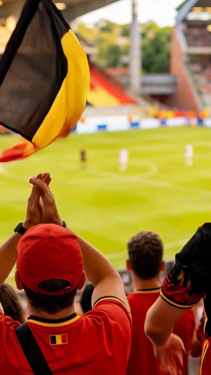 Video vertical: Aficionado central aplaudiendo con gorra roja y camiseta roja en la grada, animando el partido mientras ondea una bandera nacional.
