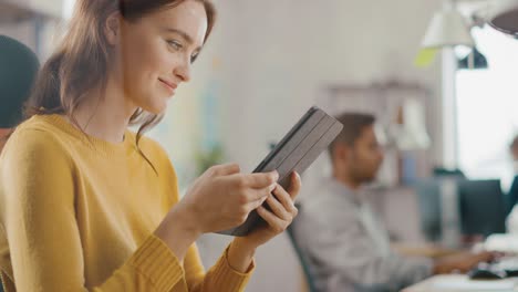 diseñadora talentosa sentada en su escritorio que está sosteniendo y usando una tableta digital de pantalla táctil. oficina brillante donde trabaja un equipo diverso de jóvenes profesionales