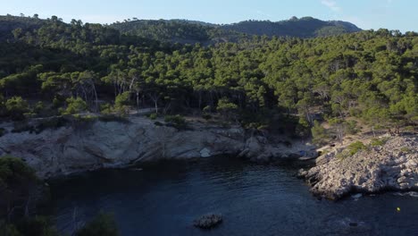Drone-flying-high-over-a-hidden-cove-in-Mallorca-spain,-the-turns-to-head-into-the-open-ocean-on-a-sunny-day