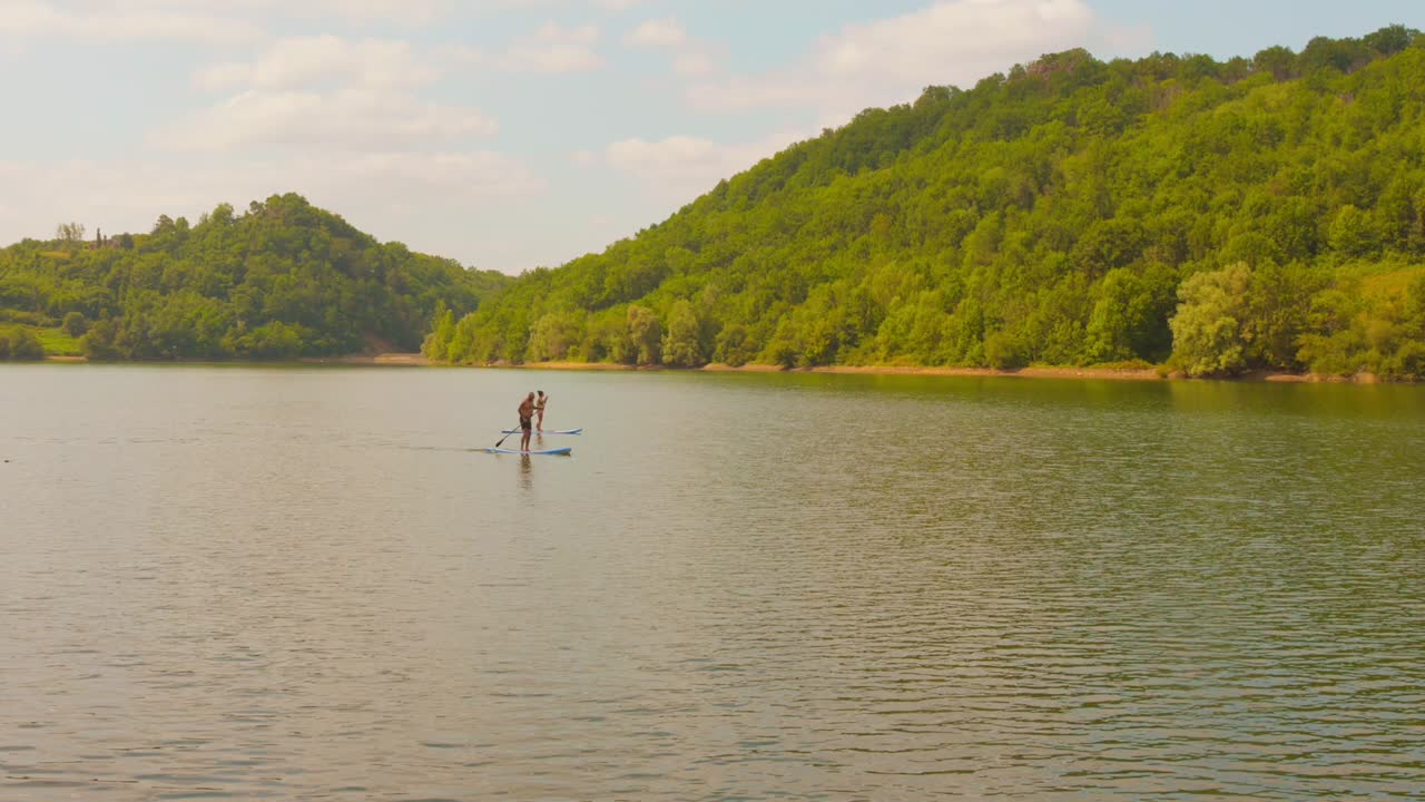 Two people enjoying paddle boarding at Lac de Mondely, France, with calm water and forested hills under a sunny sky