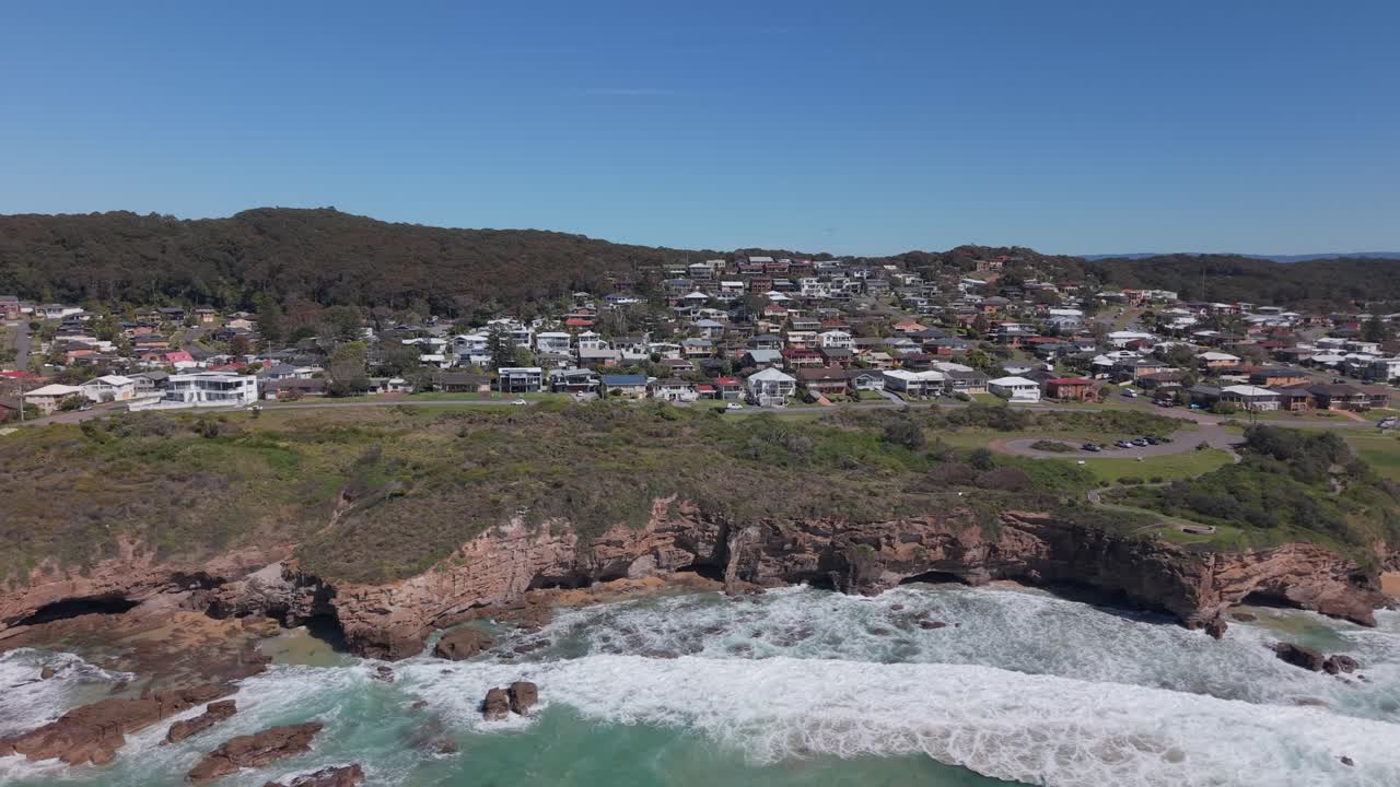 High angle drone tracking left of coastal suburb backed by trees, ocean stretches out beyond visible headland, NSW Australia