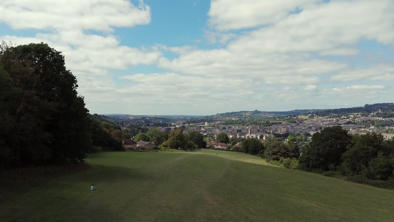 Aerial, descending, reverse shot, revealing City of Bath on a summer's day, surrounded by lush green fields with walkers and a young boy