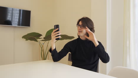 Young-man-with-glasses-waves-at-the-camera-on-his-cell-phone,-sitting-at-a-table