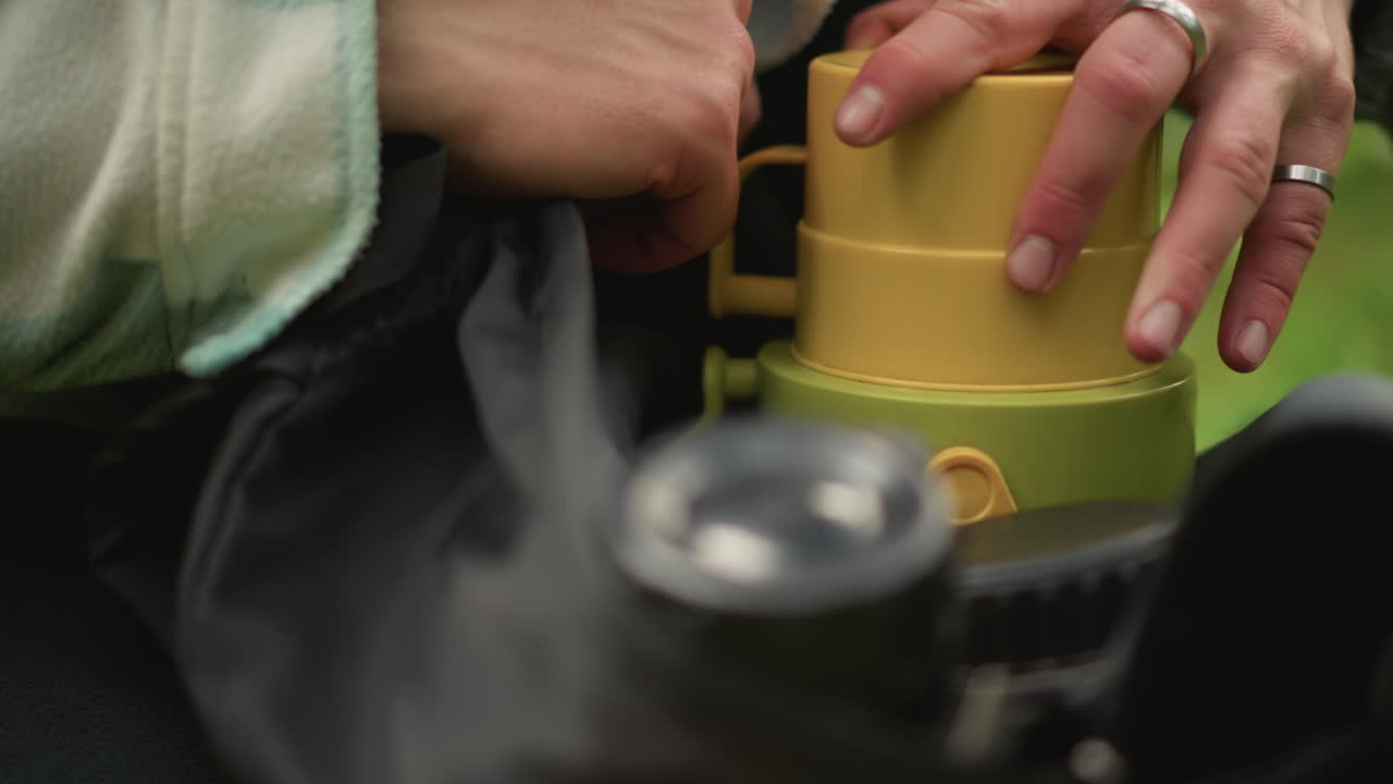 caucasian traveler checking backpack gear before hike, closeup hands adjusting thermos and camera lens, deliberate movements on damp grass, plaid jacket and ring visible, careful inspection