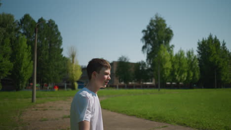 A young boy heads a ball as someone passes by in the background, with blurred trees and a building in the background