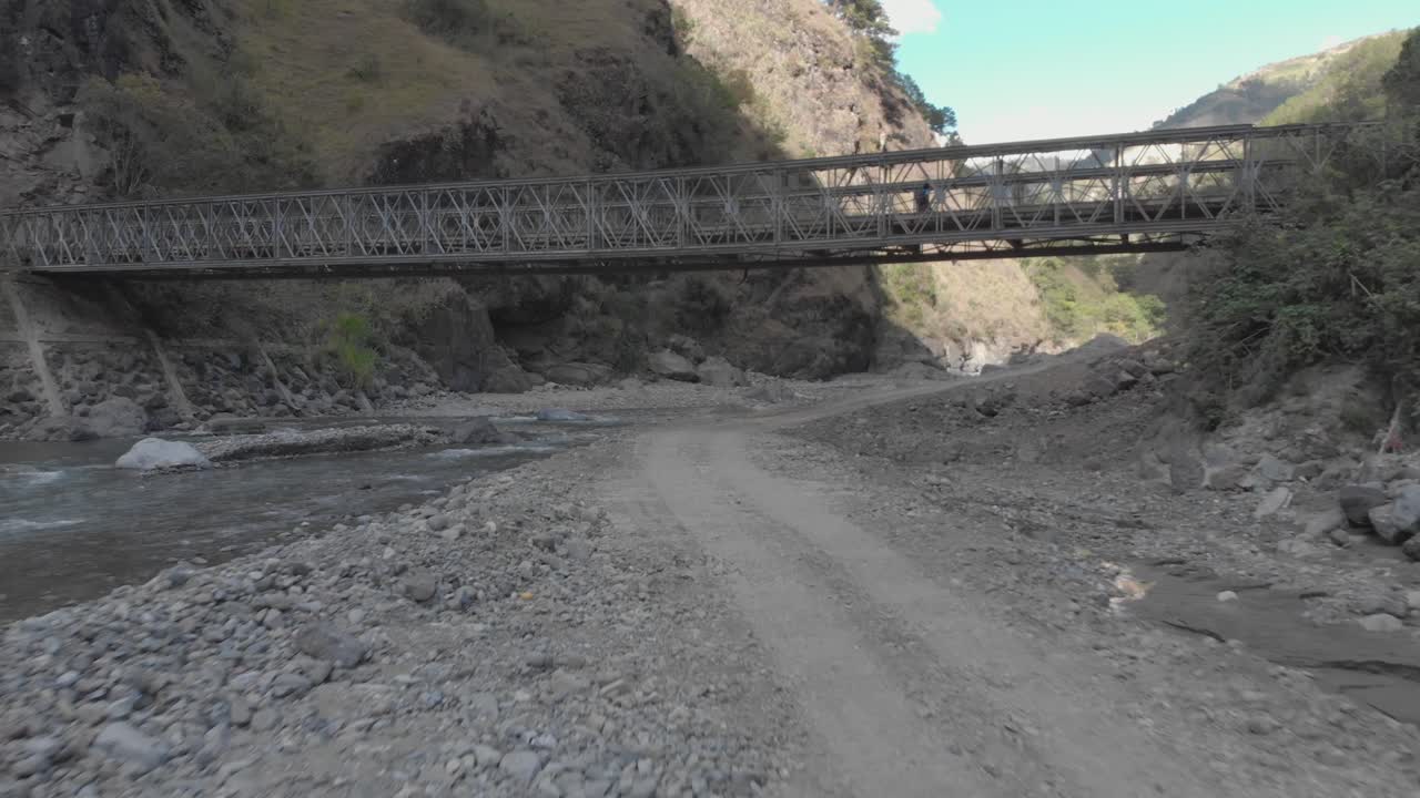 puente metálico que se extiende sobre el lecho rocoso del río en un valle montañoso rodeado de montañas árboles arbustos cepillo rocas polvo en kabayan benguet filipinas rápido vuelo bajo enfoque aéreo