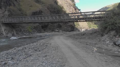 metallic bridge spanning over rocky river bed in mountainous valley surrounded by mountains trees bushes brush rocks dust in kabayan benguet philippines fast low flying aerieal approach