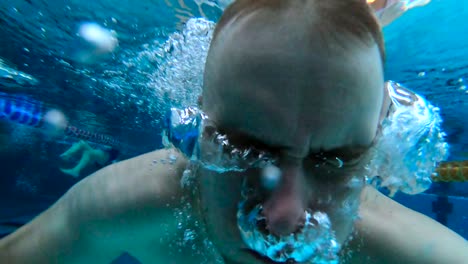 Man-is-swimming-at-an-indoor-pool-while-looking-straight-into-the-camera,-then-diving-and-blowing-air-out-of-the-nose