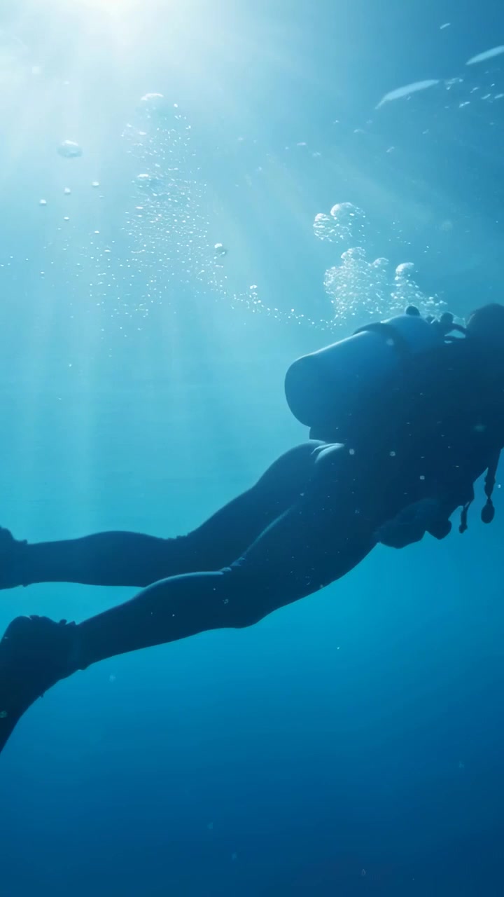 Vídeo vertical: Mergulhador com roupa de mergulho exalando bolhas, impulsionando-se em direção à superfície em oceano aberto, com cilindro.