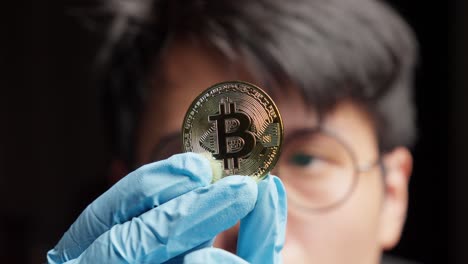 Un-Hombre-Asiático-Con-Guantes-Sostiene-Una-Moneda-Que-Representa-Btc,-Analizando-La-Tecnología