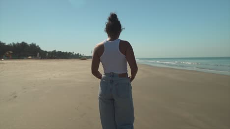 Mujer-Joven-Paseando-Por-La-Playa-En-Jeans-Y-Una-Camisa,-Cielo-Azul-Claro