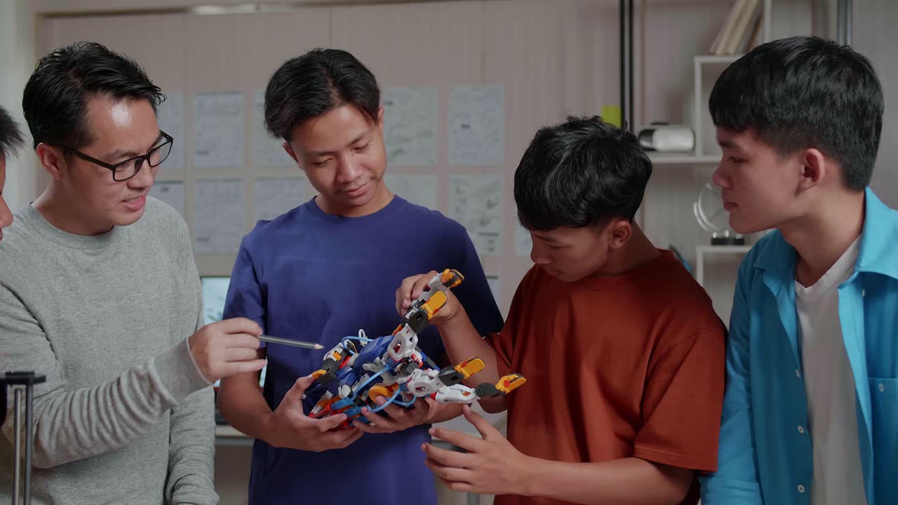 hombre asiático con una computadora portátil e impresión 3d enseñando a los niños a montar una mano de cyborg antes de celebrar el éxito en la escuela