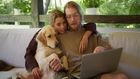 a happy family of a guy and a blonde girl, together with their dog, are considering goods in an online store on their laptop. sitting on a sofa in a gazebo in nature