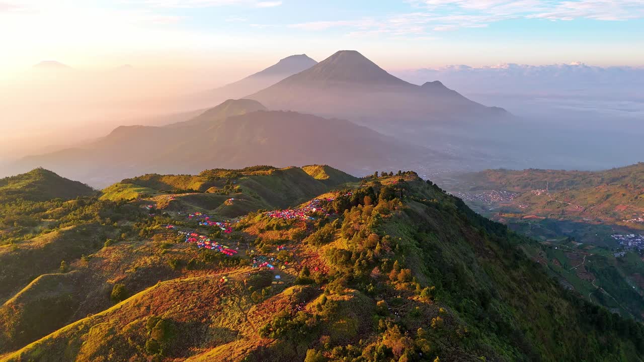 Splendida vista aerea all'alba di un campeggio di montagna con tende colorate sparse lungo la cresta e maestose montagne che si ergono sullo sfondo sopra valli nebbiose. Monte Prau, Indonesia
