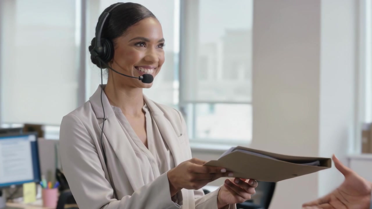 Mulher sorridente de blazer falando ao microfone, entregando um arquivo a um colega de trabalho e oferecendo o telefone no escritório para revisão.