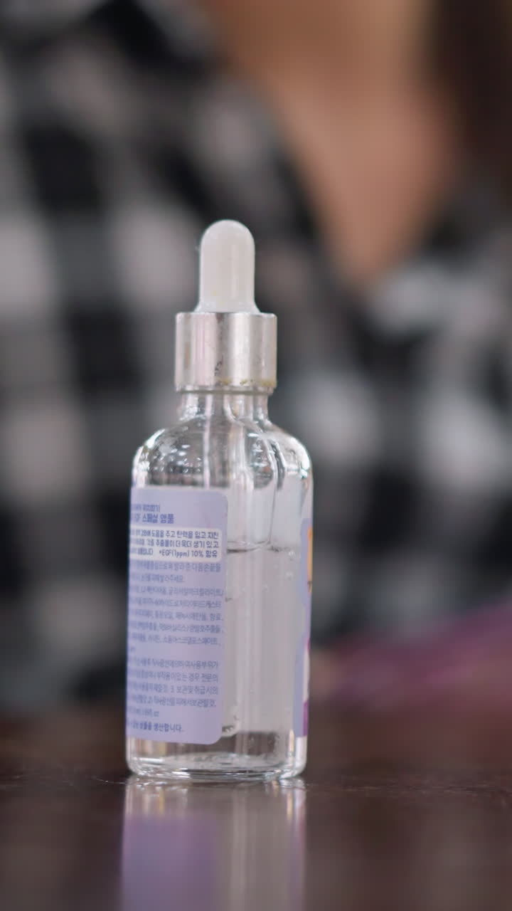 테이블 위에 놓인 스포이트 세럼 병과 파운데이션, 브러시로 제품을 바르는 체크무늬 셔츠를 입은 흐릿한 사람, 투명한 유리 피펫과 핑크색 캡, 반사되는 테이블 상판, 부드러운 보케로 연출된 편안한 홈 뷰티 장면