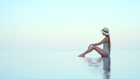 Woman-in-swimwear-and-sunhat-sitting-on-infinity-pool-border,-leaning-on-one-arm-with-a-stunning-view-of-tropical-sea-horizon-in-Bora-Bora,-static