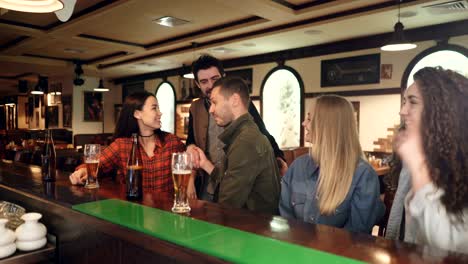 young people are talking at bar counter over beer when couple of their friends coming to meet them. men and women are greeting each other and sharing news.