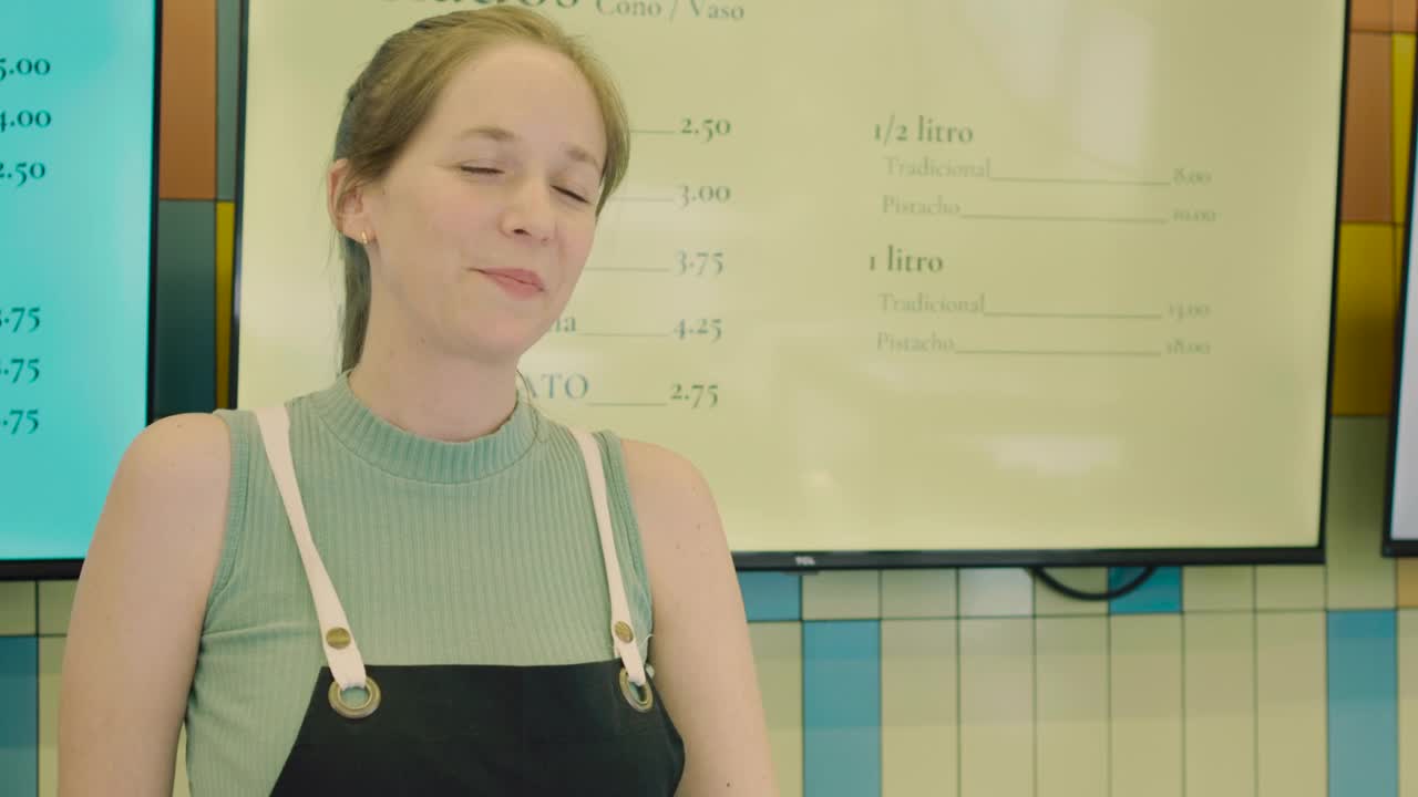 Medium shot of a young, light-eyed female ice cream shop employee handing out a sample of ice cream with a friendly and cordial expression.