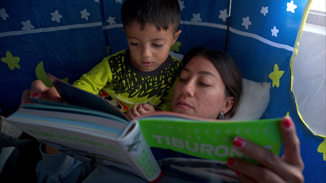 primer plano de un joven latino mexicano y su madre morena leyendo un libro acostado dentro de su tienda de castillo teniendo un momento juntos