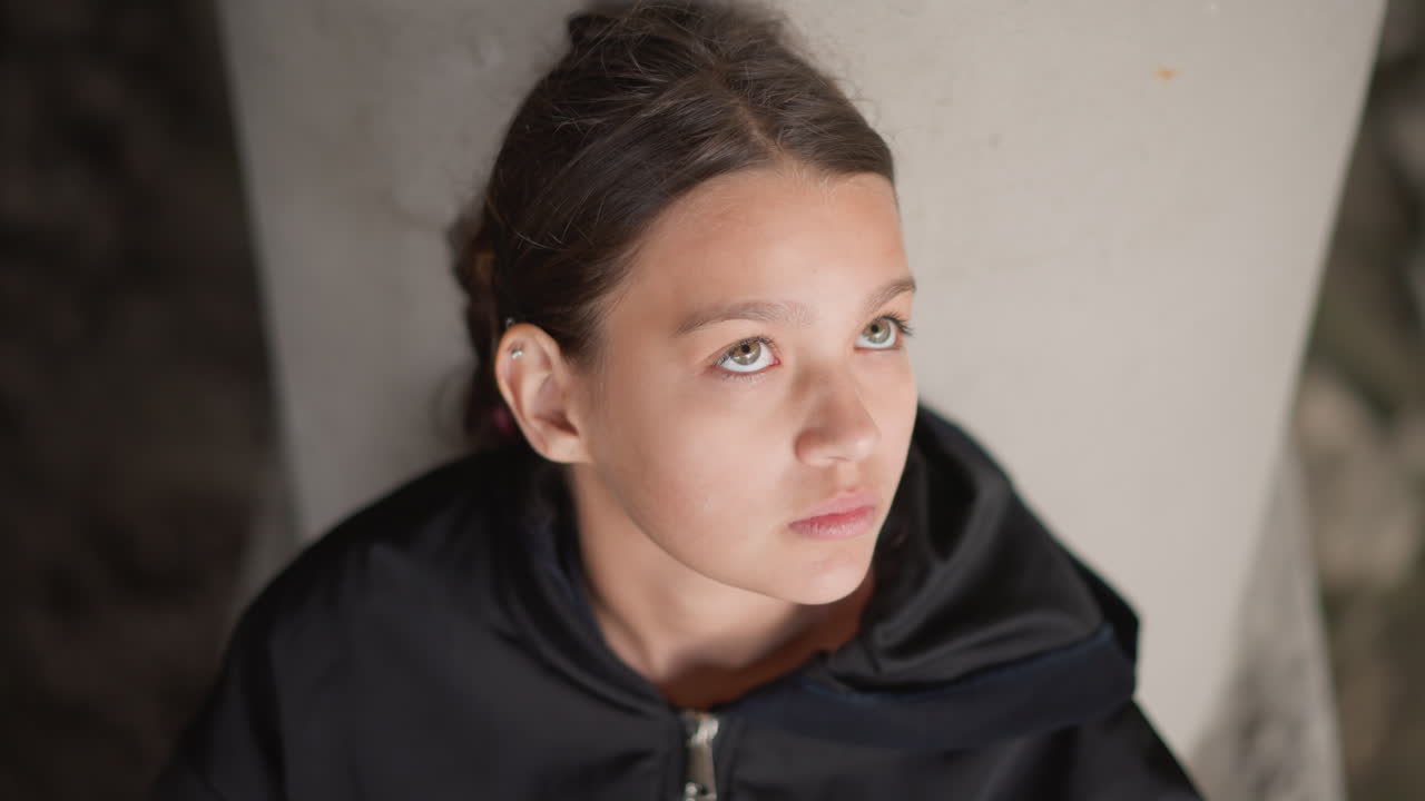 Giovane donna che guarda in alto dopo la pioggia, sguardo speranzoso verso il cielo, goccioline sui capelli, felpa con cappuccio chiusa, sfondo con colonna di cemento, delicati riflessi sulla guancia, forza tranquilla e ripresa, ritratto cinematografico