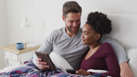 video of happy diverse couple using tablet in bed and talking