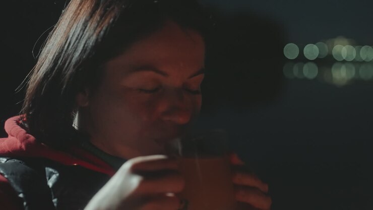 Donna bianca in campeggio in riva al lago che culla una tazza con gli occhi chiusi, luci bokeh soffuse sull'acqua, felpa con cappuccio e alito caldo, vapore che sale, umore calmo e riflessivo, atmosfera notturna della riva.