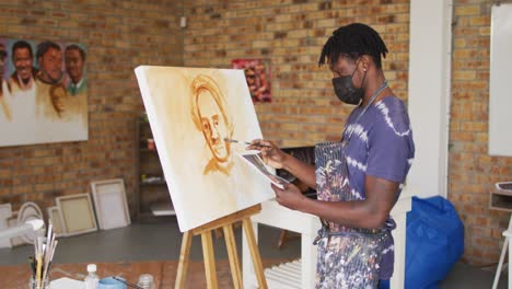 Afroamerikanischer-Männlicher-Maler-Mit-Gesichtsmaske,-Der-Im-Künstleratelier-Von-Einem-Foto-Auf-Leinwand-Malt