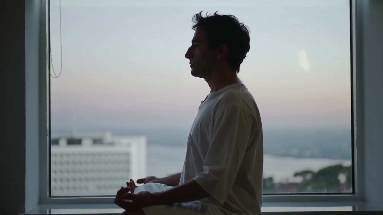 Filming man-in-white meditating on windowsill-cushion in room, seeking calm with bay-view-pull-cord