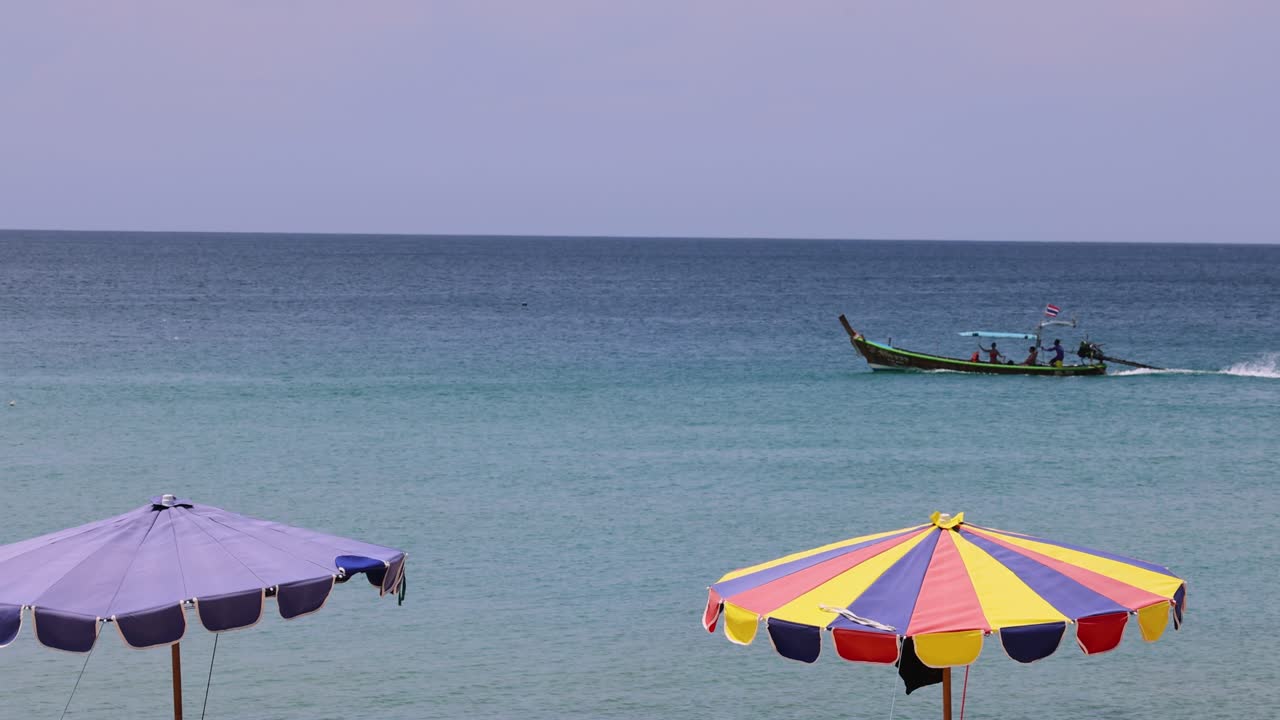 un barco de cola larga se desliza más allá de las coloridas sombrillas de playa en la playa de la libertad, phuket, bajo la suave luz del día