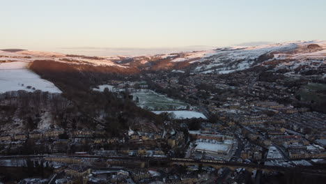 Drone-shot-of-the-winter-sun-glowing-on-the-snowy-hills-of-a-small-Yorkshire-town-with-train-going-passed