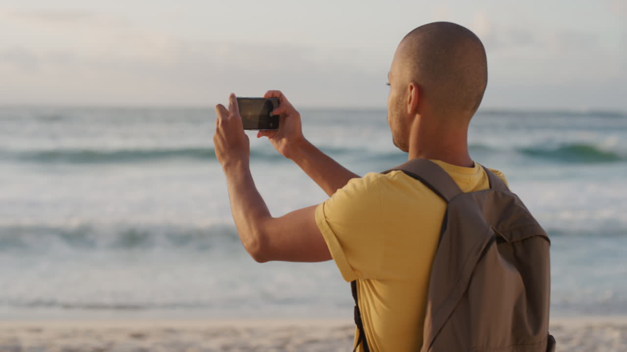 young hispanic man using smartphone taking photo of beautiful ocean seaside on beach enjoying scenic sunset view sharing vacation experience