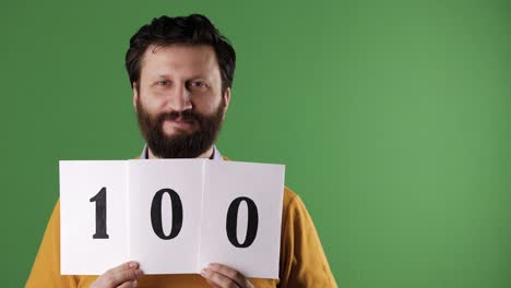 man holding number 100 sign. cheerful white bearded good-looking man in yellow sweater shows number one hundred on white sheet. chrome key, green screen, copy space