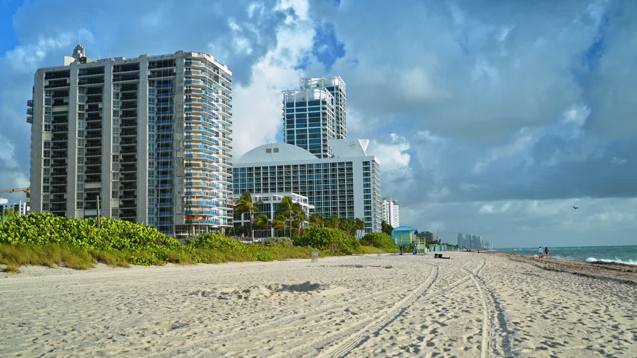 Playa de arena con huellas de neumáticos y rascacielos en Miami. Toma panorámica de la costa de North Miami Beach con huellas de vehículos en la arena y rascacielos residenciales bajo un cielo nublado.