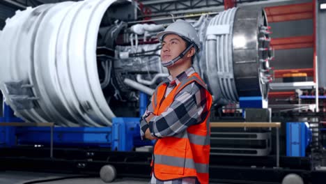 side view of asian male engineer with safety helmet crossing his arms and looking around while standing with airplane engine maintenance conducted, analytics checking the turbine