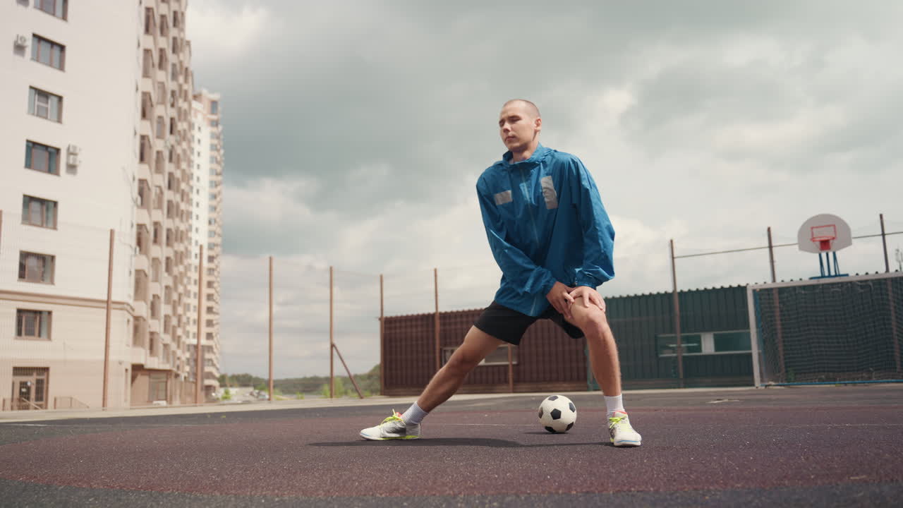 Giocatore di calcio che si allunga su un campo urbano, nuvole tempestose sopra la testa, asfalto bagnato, atleta solitario in giacca blu che esegue affondi laterali con la palla ai piedi, inquadratura dal basso, appartamenti multipiano