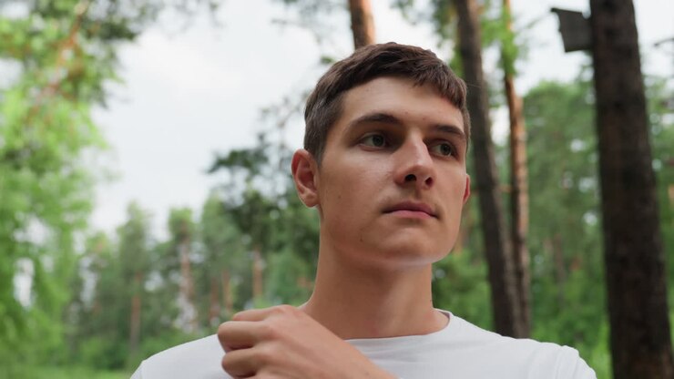 Retrato de un joven apuesto ajustándose el cuello de un polo blanco mientras está de pie al aire libre en un bosque verde, mostrando una expresión pensativa y un momento de introspección en un ambiente veraniego.