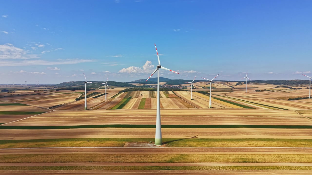 Wind turbines stand tall in an open field under a clear blue sky during daytime hours with visible farmland in the background