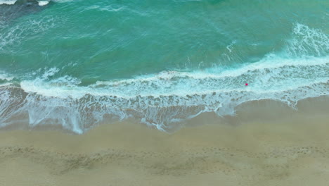 Top-down-aerial-view-of-a-beach-showing-the-foamy-waves-breaking-along-the-sandy-shore,-creating-a-natural-pattern-contrast-between-the-ocean-and-land