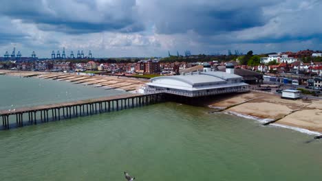 seagulls flying eyelevel and under the drone as it captures a sliding footage revealing the felixstowe pier in suffolk, its town, shipping industry, beachfront, townhouse, big rain clouds