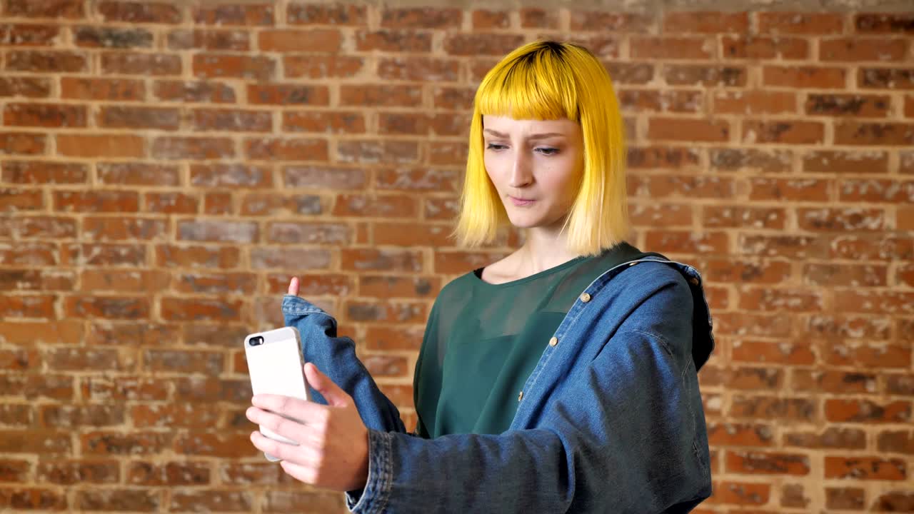 mujer joven con cabello amarillo hablando con alguien a través de chat de video, de pie cerca de fondo de ladrillo