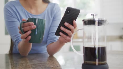 sección media de una mujer asiática sentada a la mesa, bebiendo café y usando un teléfono inteligente en la cocina