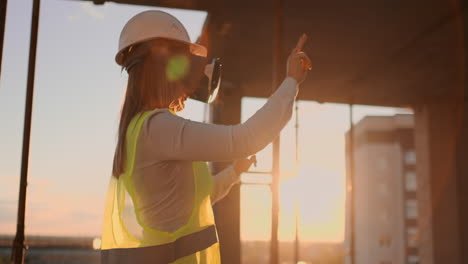 mujer ingeniera constructora en el techo del edificio al atardecer se para en gafas vr y mueve sus manos usando la interfaz del futuro. ingeniero futurista del futuro. la vista desde la parte de atrás.