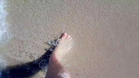 top view of man's feet walking on sandy beach barefoot splashed by sea waves