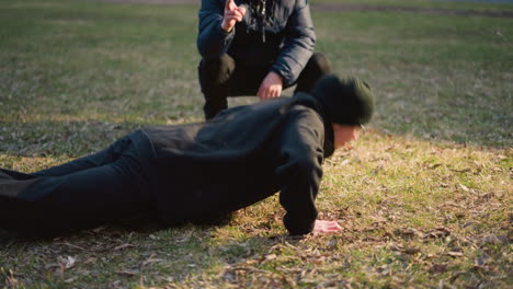 un entrenador supervisando y contando el número de flexiones realizadas por un niño vestido de negro en un campo de hierba iluminado por la luz