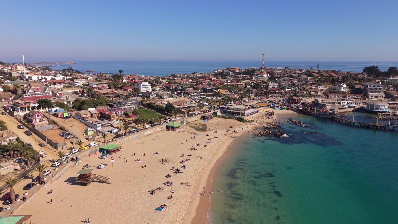 Voo avançando sobre praia de areia com banhistas e cabanas de praia, revelando a costa rochosa, o porto e o horizonte denso da cidade costeira. El Quisco, Chile