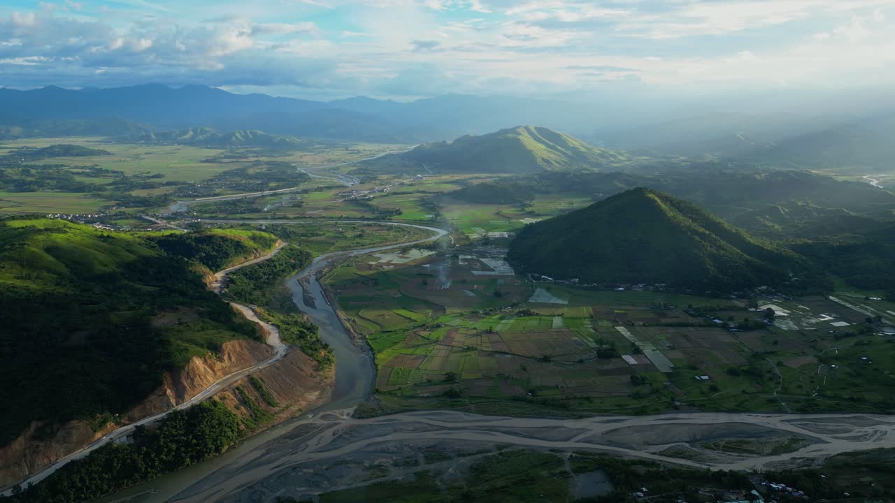 A high pan of Bagabag reveals the Santa Cruz River winding through fields with sunbeams lighting mountain peaks Bambang, Nueva Vizcaya, Philippines